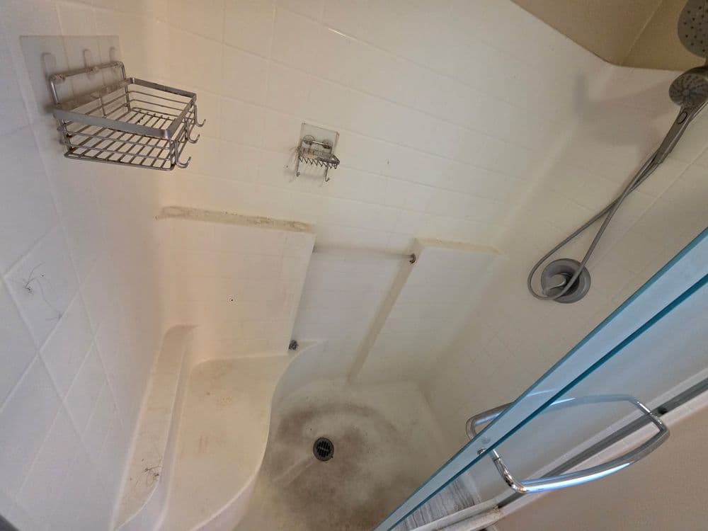 Empty shower stall with white tiled walls, grab bars, and a drain, showing signs of wear.